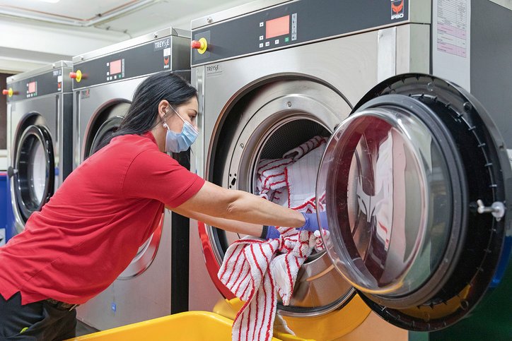 Eine Frau mit langen Haaren und einer Maske wirft Wäsche in eine Industriewaschmaschine. Sie befindet sich in einem Raum voller Waschmaschinen.