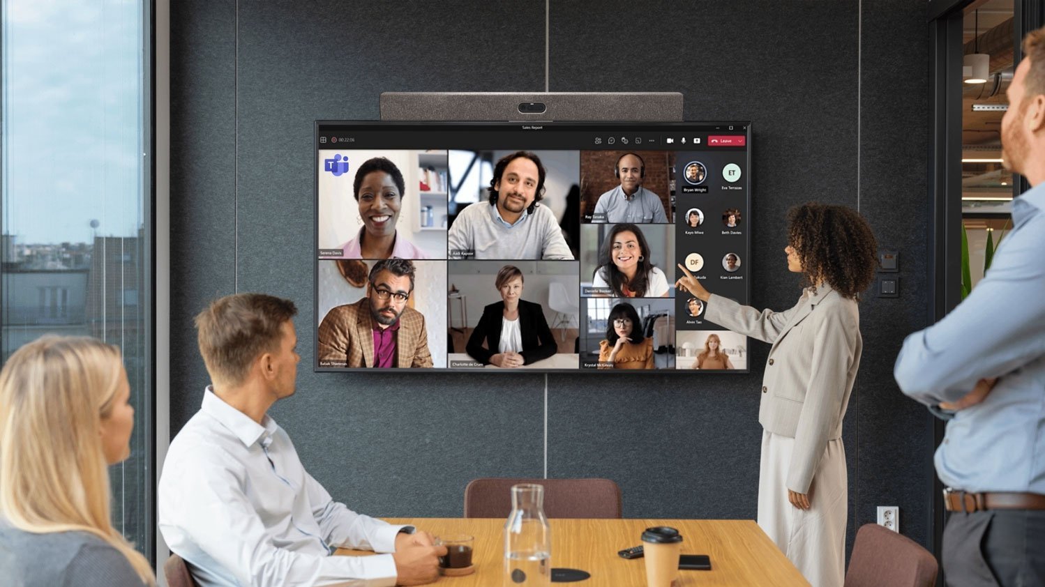 Eine Gruppe Menschen steht vor einem Bildschirm, auf welchem ein Teams-Meeting angezeigt wird.