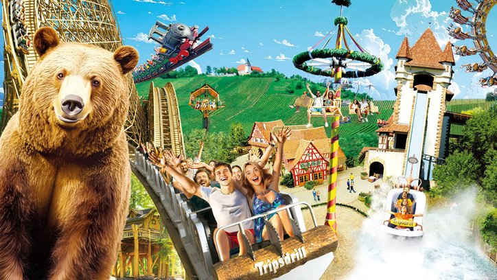 Auf dem Bild sind verschiedene Achterbahnen zu sehen, in denen Leute sitzen, die Spaß haben. Im Vordergrund ist ein großer Bär zu sehen. 