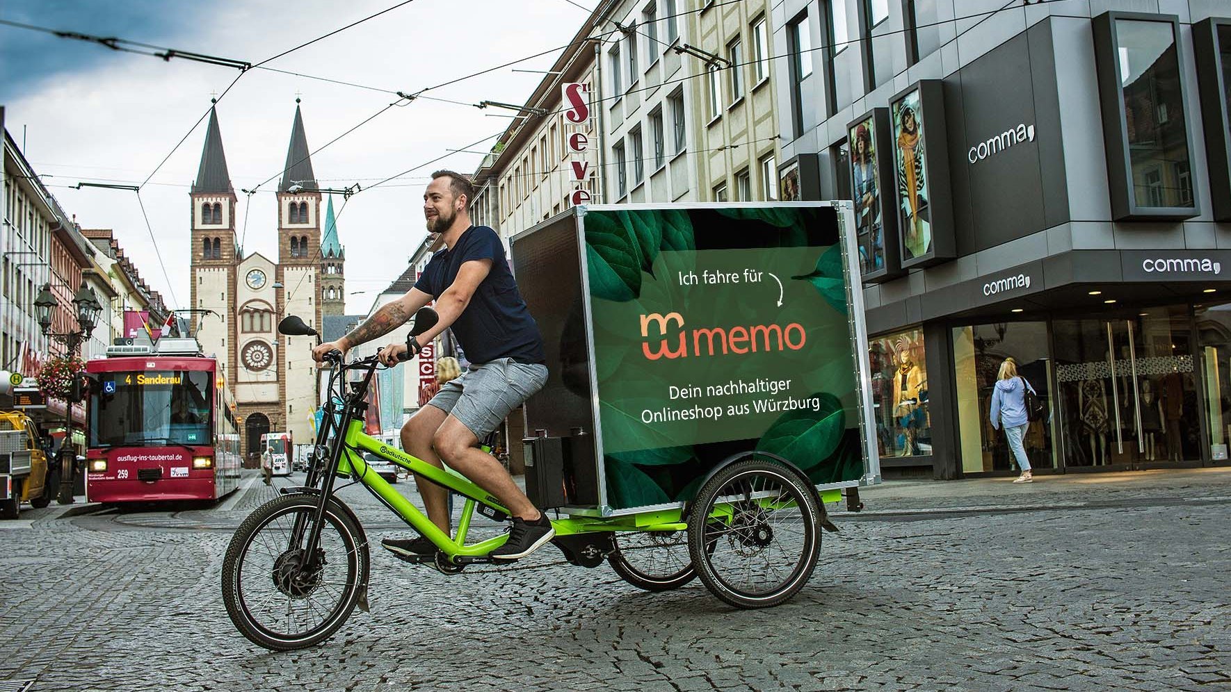 Ein Kurier fährt mit einem grünen Lastenrad durch eine Innenstadt. Auf dem Transportbehälter steht: „Ich fahre für memo – Dein nachhaltiger Onlineshop aus Würzburg“. Im Hintergrund sind eine Straßenbahn, historische Gebäude und Türme zu sehen.