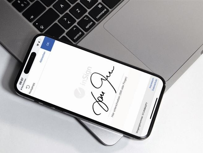 Das Bild zeigt ein Smartphone, welches auf einem Laptop liegt. Auf dem Bildschirm ist eine digitale Unterschrift zu sehen.
