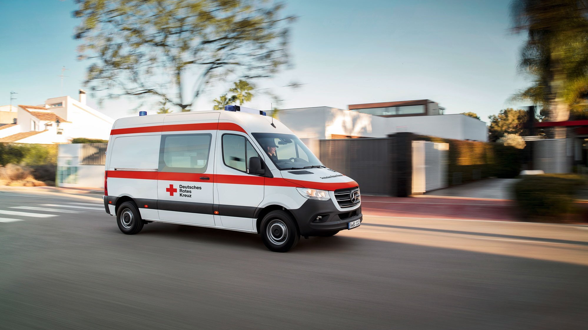 Ein Rettungswagen fährt mit eingeschaltetem Blaulicht durch ein städtisches Wohngebiet. Der weiße Mercedes-Benz Sprinter hat auf der Seite gut sichtbar das Rote Kreuz Logo und den Schriftzug „Deutsches Rotes Kreuz“ platziert. Durch die Bewegungsunschärfe im Hintergrund wirkt die Szene dynamisch.