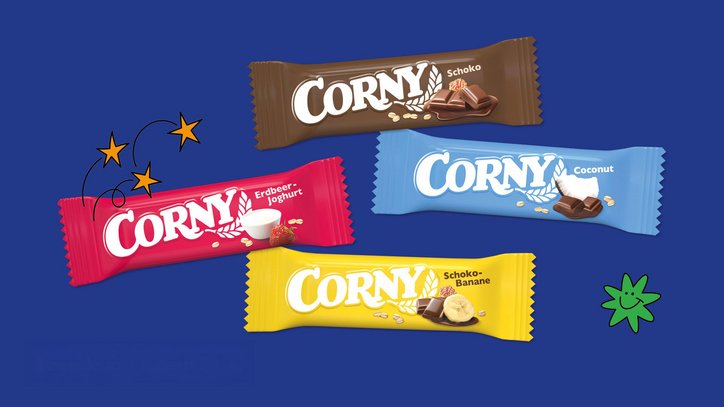 Vier CORNY-Müsliriegel in unterschiedlichen Geschmacksrichtungen auf blauem Hintergrund. Die Verpackungen sind farbig gestaltet und zeigen Sorten wie Schoko, Coconut, Erdbeer-Joghurt und Schoko-Banane.
